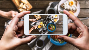 Hand taking photo of top view shot breakfast table with mobile phone. Poached egg with bread, sliced orange and avocado are on plates. Coffee mugs, cheese plate and salad with avocado, tomatoes and arugula are on the table with a grey tea towel. Hand slicing egg on the plate.